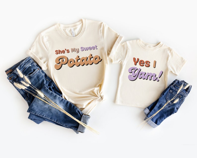 Pu&ograve; includere: Due magliette color crema con le frasi "She's My Sweet Potato" e "Yes I Yam!" in lettering in stile retr&ograve;. Le magliette sono abbinate a jeans blu ed erba secca decorativa, suggerendo un outfit coordinato per una famiglia o una coppia.