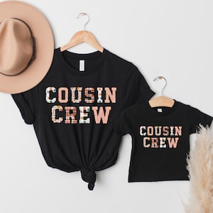Puede incluir: Dos camisetas negras, una para adultos y otra para niños, colgadas de perchas de madera. Las camisetas muestran el texto "COUSIN CREW" en un diseño estampado en rosa, blanco y marrón. Un sombrero color canela es visible en la esquina superior izquierda.