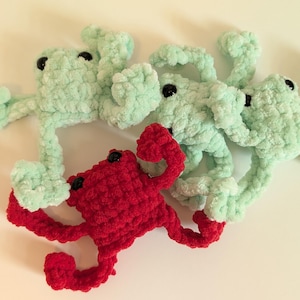 May include: Four crocheted frog toys. Three are light green, and one is red. Each frog has black button eyes and outstretched limbs, giving them a playful appearance. The frogs are arranged on a light-colored surface.