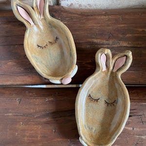 Könnte beinhalten: Zwei Keramik-Schalen in Hasenform mit geschlossenen Augen und rosa Ohren. Die Schalen sind in Braun und Beige glasiert und haben eine gesprenkelte Oberfläche.