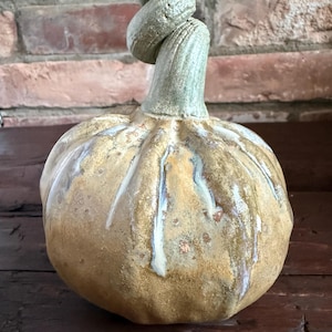 Könnte beinhalten: Eine Kürbisskulptur aus Keramik mit einem abgerundeten Körper und einer texturierten, gesprenkelten Glasur in Beige und Braun. Der Kürbis hat tiefe vertikale Rillen und einen markanten, geschwungenen grünen Stiel an der Oberseite.