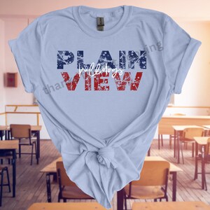 Peut inclure: T-shirt bleu clair avec les mots "PLAIN VIEW" en lettres rouges et bleues. Le mot "bulldogs" est écrit en cursive entre les mots "PLAIN" et "VIEW". Le t-shirt est noué en bas.