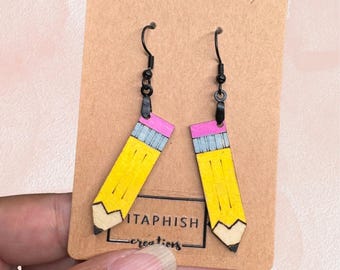May include: A pair of handmade pencil earrings. Each earring is a yellow pencil design with a pink eraser, blue metal band, and a black hook. The earrings are displayed on a brown card with the text "ITAPHISH creations."