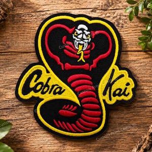 Pode incluir: Um emblema bordado apresentando uma cobra estilizada com corpo preto, detalhes em vermelho e uma língua amarela. O texto Cobra Kai está bordado em letras pretas sobre um fundo amarelo, com uma borda amarela espessa contornando o design.