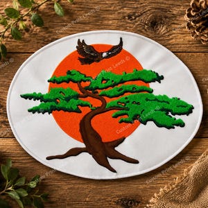 May include: An oval-shaped fabric patch featuring an embroidered design of a brown bonsai tree with vibrant green foliage set against a large, solid orange circular background. A small black eagle is depicted flying above the tree.