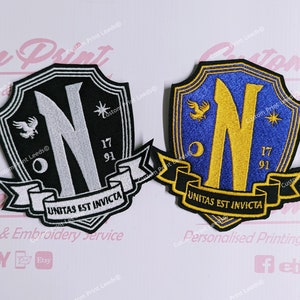 May include: Two embroidered patches featuring a black shield shape, a white letter N, a white bird, a white star, and a white crescent moon. The text on the banner reads "UNITAS EST INVICTA". The other patch has a blue shield shape, a gold letter N, a gold bird, a gold star, and a gold crescent moon. The text on the banner reads "UNITAS EST INVICTA".