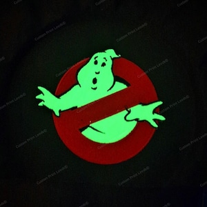 May include: A black garment featuring a glowing light green ghost graphic centred within a red circular emblem with a diagonal slash. The design is surrounded by a repeating watermark text that reads Custom Print Leeds across the dark background.
