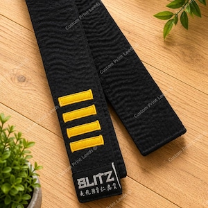 May include: A black martial arts belt featuring a textured, wavy fabric surface. The belt includes four horizontal yellow embroidered stripes and a black rectangular patch with the white text BLITZ and Japanese characters at the end.