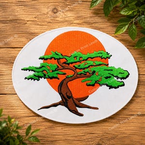 Può includere: Una toppa circolare in tessuto bianco con il disegno ricamato di un bonsai marrone con fogliame verde brillante. Dietro l'albero si trova un grande cerchio arancione pieno. La toppa presenta un bordo cucito netto e una forma circolare.