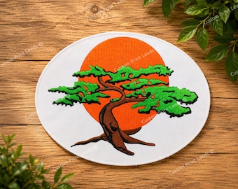 May include: A circular white fabric patch featuring an embroidered design of a brown bonsai tree with vibrant green foliage. Behind the tree is a large, solid orange circle. The patch has a clean, stitched border and a flat, circular shape.