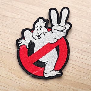 Può includere: Una toppa ricamata raffigurante un personaggio fantasma bianco con un'espressione sorridente, che fa il segno di pace con una mano. Il fantasma è posizionato davanti a un simbolo circolare rosso con una linea diagonale rossa, bordato in nero.