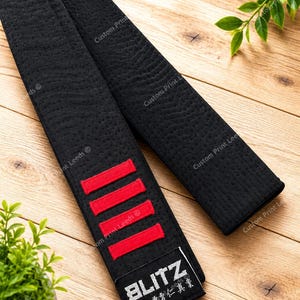 May include: A black martial arts belt featuring a textured fabric surface, four horizontal red embroidered stripes, and a black rectangular patch at the end. The patch displays the white text BLITZ above a row of Japanese kanji characters.