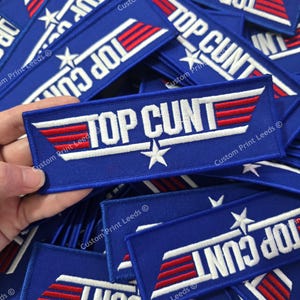 May include: A rectangular blue embroidered patch featuring the white text TOP CUNT with a white star below the word CUNT. The design includes stylized red and white horizontal stripes extending from the letters, mimicking a classic aviation emblem.