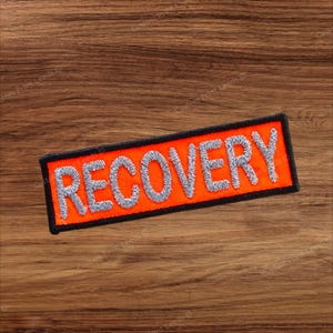 Könnte beinhalten: Ein rechteckiger, bestickter Aufnäher mit leuchtend orangefarbenem Hintergrund und einem dicken schwarzen Rand. Das Wort RECOVERY ist in großen, hellgrauen Großbuchstaben in die Mitte des Aufnähers gestickt, was ein kontrastreiches Design ergibt.