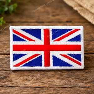 May include: A rectangular embroidered patch featuring the Union Jack flag design. The patch displays a central red cross with white borders, set against a blue background with diagonal red and white stripes, all finished with a white stitched border.