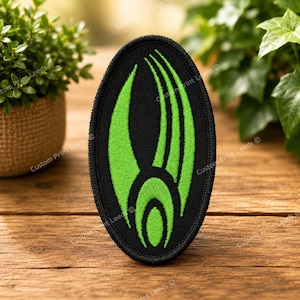 Peut inclure: Un écusson brodé de forme ovale présentant un fond noir avec un motif graphique vert néon vif. Le design se compose d'une forme ovale centrale flanquée de trois lignes courbes sur la droite et d'une grande forme courbe sur la gauche.
