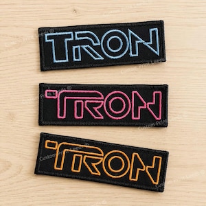 May include: Three rectangular embroidered patches featuring the word TRON in a stylized font. The top patch has light blue lettering on a black background, the middle patch has pink lettering, and the bottom patch has orange lettering.