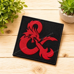 May include: A square black fabric patch featuring a red embroidered dragon design in the shape of an ampersand. The patch has a stitched border and a flat, textured surface, showcasing a detailed mythical creature motif in bold red thread.