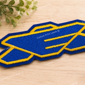 May include: An embroidered patch featuring a geometric design in royal blue with a thick, bright yellow border. The shape consists of two overlapping, elongated hexagonal forms that create a stylized, angular pattern with sharp, pointed edges.
