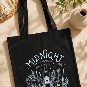 Puede incluir: Un bolso tote negro con un estampado gráfico blanco que incluye el texto Midnight Margaritas. El diseño muestra una botella de tequila, una copa de margarita, un mortero con mano, polillas y varios elementos botánicos en el frente.