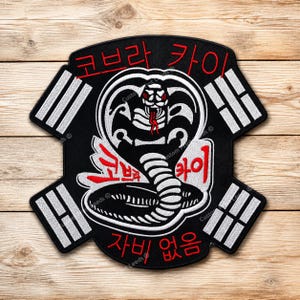 Op de afbeelding: Een zwarte geborduurde patch met een centrale witte cobra-slangafbeelding met rode accenten. Het ontwerp bevat Koreaanse tekst in rood en wit, omringd door vier witte balksymbolen in de hoeken, die doen denken aan een vlag-achtig embleem.