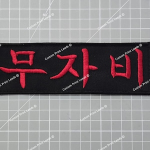 May include: A black rectangular patch featuring red Japanese characters. The embroidered text reads "무자비". The patch is set against a grey grid background. The words "Custom Print Leeds" are printed around the edges.
