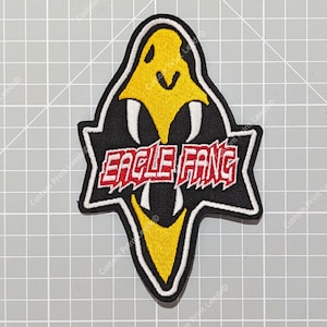 Puede incluir: Un parche bordado negro y amarillo con las palabras "Eagle Fang" en letras rojas y blancas. El parche presenta una cabeza de águila estilizada con colmillos.