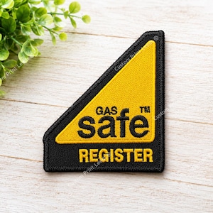 May include: A triangular embroidered patch with a black border and a yellow centre. The patch features the black text GAS safe TM and the yellow text REGISTER below it. The design is oriented with a diagonal edge on the top right side.