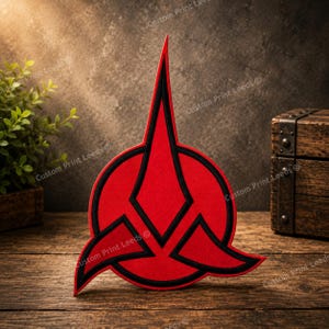 May include: A red and black emblem featuring a circular base with a tall, pointed vertical spike and two curved side wings. The design is flat with bold black outlines separating the red sections, creating a sharp, symmetrical, and stylized shape.