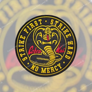 May include: A round black patch featuring a yellow cobra emblem and text. The text, in yellow, reads "STRIKE FIRST, STRIKE HARD, NO MERCY". The cobra has a red tongue and the words "Cobra Kai" are in red.