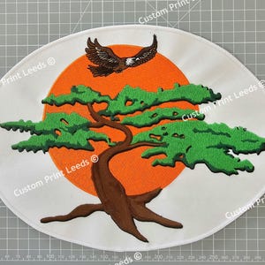 May include: Embroidered patch featuring a green bonsai tree with a brown trunk against an orange sun. The patch has white fabric with the text "Custom Print Leeds" repeated around the edge.