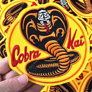 Peut inclure: Un écusson brodé circulaire représentant un cobra noir et orange avec une tête blanche et une langue rouge. Le fond est jaune vif avec une bordure rouge. Le texte Cobra Kai est brodé en écriture rouge sur les côtés gauche et droit.