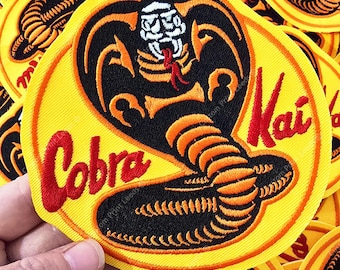May include: A circular embroidered patch featuring a black and orange cobra snake with a white head and red tongue. The background is bright yellow with a red border. The text Cobra Kai is embroidered in red script on the left and right sides.