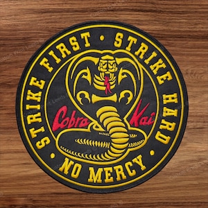 Puede incluir: Un parche bordado circular con fondo negro y un gráfico de cobra amarilla en el centro. El texto dice Strike First, Strike Hard, No Mercy en amarillo, con el texto en rojo Cobra Kai sobre el cuerpo de la serpiente.
