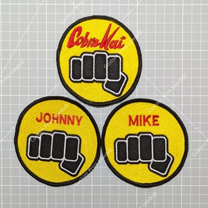 Puede incluir: Tres parches bordados circulares. El superior muestra "Cobra Kai" en rojo sobre fondo amarillo. Los otros dos presentan un diseño de puño negro y los nombres "JOHNNY" y "MIKE" en rojo, todo sobre fondo amarillo.