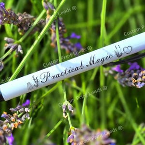 May include: A white pen with the words "Practical Magic" in script, along with small illustrations of a mortar and pestle, wheat, and a heart. The pen is set against a backdrop of lavender flowers and green foliage.