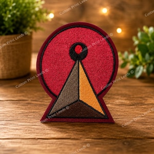 May include: A circular embroidered patch featuring a deep red background with a black border. Centered is a black circular icon above a three-dimensional pyramid shape divided into three distinct sections coloured in dark brown, light brown, and gold.