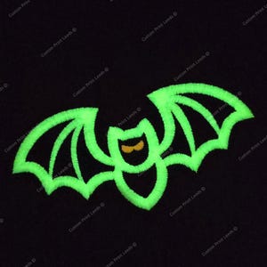 Puede incluir: Un diseño de murciélago bordado en verde neón sobre un fondo negro. El murciélago tiene las alas extendidas y una pequeña cara de color naranja. El diseño es probablemente para Halloween o un tema similar.