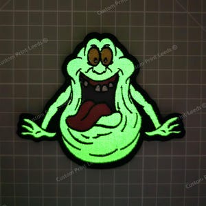 May include: A green glow-in-the-dark embroidered patch of a cartoon ghost with a wide open mouth and tongue sticking out. The ghost has two eyes and a big smile.