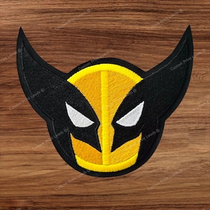 May include: An embroidered patch featuring a stylized mask design in black and yellow thread. The mask has two large, pointed black ears, a central yellow vertical stripe, and two white triangular eye cutouts. The edges are finished with black stitching.