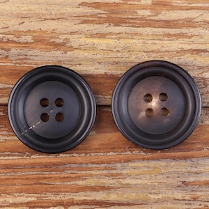 Peut inclure: Deux gros boutons noirs ronds avec quatre trous chacun. Les boutons ont un bord légèrement surélevé et reposent sur une surface en bois vieilli. Un bouton a une petite rayure blanche.