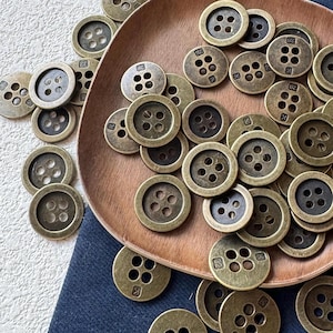May include: A collection of antique brass-colored buttons with four holes, some with the number "32" or the letter "K" stamped on them. The buttons are scattered on a wooden plate and a dark blue fabric.