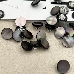 Peut inclure: Une collection de boutons ronds de différentes tailles et nuances de noir, de gris et de nacre irisée. Certains boutons ont une surface brillante et réfléchissante, tandis que d'autres sont mats. Les boutons sont dispersés sur une surface beige clair.