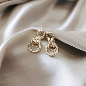 May include: A pair of gold-toned earrings with a linked chain design. The earrings are on a light brown fabric background.