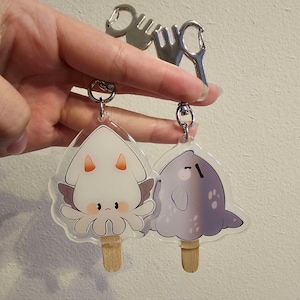 Könnte beinhalten: Zwei transparente Acryl-Schlüsselanhänger in Form von Cartoon-Tintenfisch- und Seehund-Eis am Stiel. Der Tintenfisch ist weiß mit orangefarbenen Akzenten und der Seehund ist lila. Jeder Schlüsselanhänger hat einen silbernen Ring und einen Holzstab.