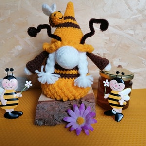May include: A crocheted gnome dressed as a bee with brown and yellow stripes, holding a small bee on its hat. The gnome is sitting on a wooden log with a purple flower in front of it. There are two bee figurines on either side of the gnome, one holding a flower and the other next to a jar of honey.