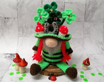 Op de afbeelding: Een groene gehaakte gnome met een hoed met een hoefijzer en klavertjesvier. De gnome zit op een houten basis met rode paddenstoelen en groene klavertjesvier eromheen.