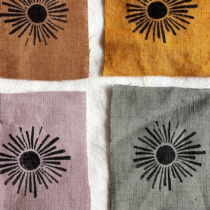 May include: Four fabric squares with a black sunburst design printed on each. The squares are in four different colours: brown, yellow, purple, and grey.