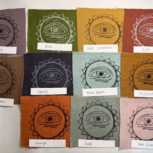 Può includere: Campioni di tessuto in vari colori, ciascuno con un disegno di occhio in bianco e nero all'interno di un sole. I colori includono lavanda, oliva, cannella chiara, ruggine, blu navy, verde menta, senape, vero cannella, arancione, salvia e rosa pallido.