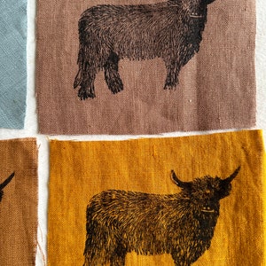 May include: A black and white illustration of a Highland cow printed on a brown linen fabric. The fabric is textured and has a natural look.